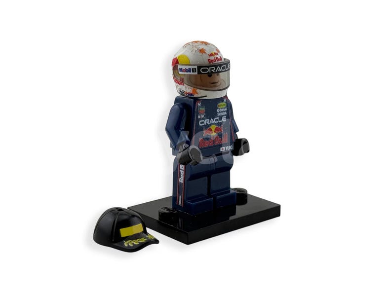 May include: A Lego figurine of a race car driver wearing a helmet with the Mobil 1 and Oracle logos. The figurine is in a dark blue racing suit with the Red Bull logo. A black baseball cap with a yellow design sits next to the figurine.