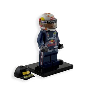 May include: A Lego figurine of a race car driver wearing a helmet with Mobil 1 and Oracle branding. The figurine is dressed in a dark blue racing suit with Red Bull and Oracle logos. A black baseball cap with gold accents sits nearby.