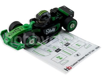 May include: A black and green toy race car with the number 27 and sponsor logos. The car has a driver figure wearing a helmet. A sheet of custom stickers is next to the car.