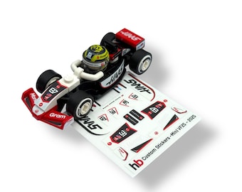 May include: A red, black, and white toy race car with a driver figure. The car has the number 87 and the word "HAAS" on it. A sheet of custom stickers is next to the car, with the text "Custom Stickers - Mini VF25 - 2025".