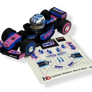 May include: A small black, blue, and pink race car model with a driver's helmet. The car features the number 10 and BWT branding. Beside it is a sheet of custom stickers, with the text "hb Custom Stickers Mini A A524 - 2024".