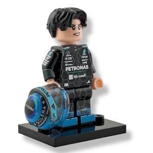 May include: A Lego figurine of a race car driver in a black and teal racing suit, holding a matching helmet. The helmet has the words "SOLERA" and "Meta A" printed on it. The figurine is standing on a black base.