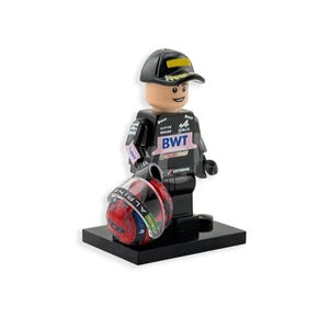 Op de afbeelding: Een Lego-figuur van een autocoureur in een zwart uniform met sponsorlogo's, een zwarte pet en een rood-blauwe helm. Het figuur staat op een zwarte basis.