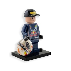 May include: A Lego figurine of a race car driver wearing a navy blue uniform with the Oracle logo and a black cap. The figurine is holding a white and orange helmet with the Oracle logo and is standing on a black base.