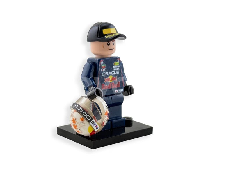 May include: A Lego figurine of a race car driver wearing a navy blue uniform with the Oracle logo and a black cap. The figurine is holding a white and orange helmet with the Oracle logo and is standing on a black base.