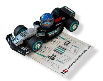 May include: A black and teal Lego race car with a driver in a blue helmet. The car sits on a sheet of stickers with the text "Custom Stickers Mini M W16-2025". The car has the word "PETRONAS" on it.