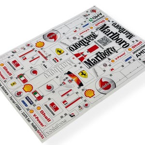 以下が含まれることがあります： フェラーリF2004モデル用の代替ステッカーシート。さまざまなロゴとブランドが特徴です。「Marlboro」ロゴは白と赤で、他に「Shell」、「Vodafone」などのスポンサーロゴがあります。シートは白で光沢があります。