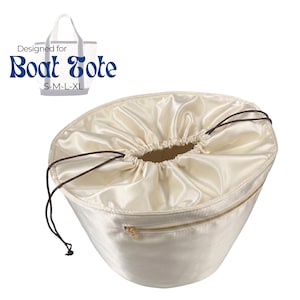 Könnte beinhalten: Cremefarbene Boat Tote-Tasche aus Satin mit Kordelzug und goldenem Reißverschluss. Die Tasche hat eine abgerundete Form und einen braunen Kordelzug. Der Text "Designed for Boat Tote" befindet sich oben links.