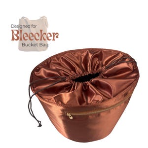 May include: A brown satin bucket bag with a drawstring closure and a gold zipper. The bag is designed for the Bleecker Bucket Bag. The text "Designed for Bleecker Bucket Bag" is printed on the top left.