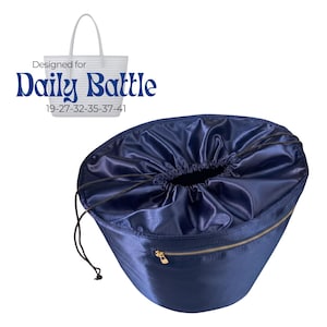 May include: A navy blue drawstring bag with a gold zipper and the text "Designed for Daily Battle". The bag has a shiny, satin-like texture and a drawstring closure. A white tote bag is in the background.