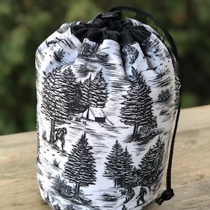 May include: A white drawstring bag with a black and white print of trees, a tent, and a Bigfoot. The bag has a black drawstring.