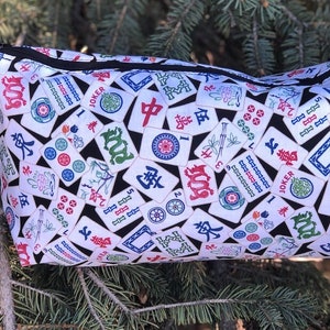 May include: A white zippered pouch with a black background and a pattern of colorful Mahjong tiles. The tiles feature Chinese characters, numbers, and symbols.