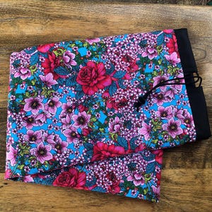 May include: A fabric item with a floral pattern, showcasing red roses and pink flowers on a bright blue backdrop. It has a black border and a drawstring closure.