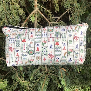 May include: Rectangular white zippered pouch with a repeating pattern of Mahjong tiles in red, green, blue, and black. The pouch is set against a backdrop of green pine needles and branches.
