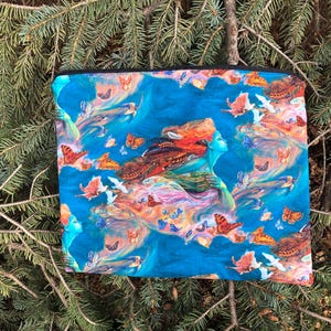 May include: A rectangular, zippered pouch featuring a vibrant, artistic design. The artwork depicts a woman with flowing red hair and blue skin, surrounded by butterflies and birds against a blue background. The pouch is set against a backdrop of evergreen branches.