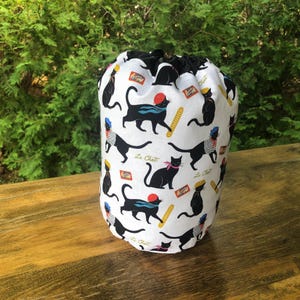 May include: A white drawstring bag with a black and white cat pattern. The cats are depicted in various poses, some wearing berets and scarves. The bag also features the text "Le Chat" and images of baguettes. The top of the bag is black.