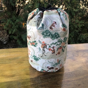 May include: A cylindrical drawstring bag with a whimsical print featuring frogs, mice, and vegetables in a garden scene. The bag's fabric is primarily white with green, brown, and orange accents. The top of the bag is black.