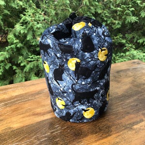 May include: A cylindrical fabric bag with a drawstring closure. The bag features a dark blue background with a pattern of black cats, yellow moons, and bare tree branches. The bag is sitting on a wooden surface.