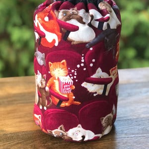 May include: A red drawstring bag featuring a pattern of cats seated in cinema chairs. Some cats are holding popcorn boxes labelled "Kitty Corn".