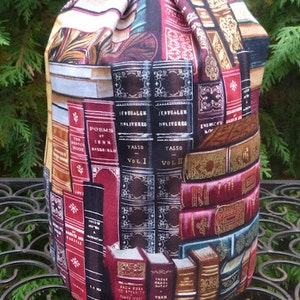 May include: A cylindrical drawstring bag with a repeating pattern of antique books in various colors. The books have titles like "Tasso Vol. I" and "Poems of John Keats". The bag is made of fabric and has a black drawstring closure.