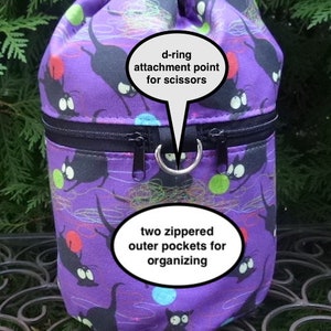 May include: Purple drawstring bag with a black cat print and two zippered outer pockets. The bag is 9 inches tall and 5.5 inches in diameter. It has a d-ring attachment point for scissors.  "stock image" is written at the bottom of the image.