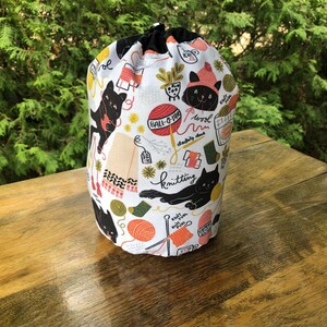 May include: A cylindrical fabric project bag with a drawstring closure, featuring a white background printed with black cats, colorful yarn balls, knitting needles, and text including "BALL-O-YARN", "knitting", "knit", and "purl" in various fonts.