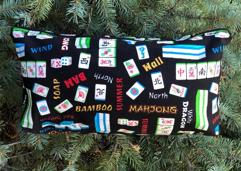 Puede incluir: Bolsa rectangular negra con cremallera y un estampado de fichas de Mahjong. La bolsa presenta varias fichas de Mahjong, dados y texto como "BAM", "North" y "Mahjong". El fondo es negro y el ribete es verde.