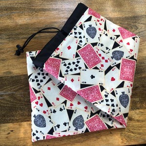 May include: A fabric drawstring pouch featuring a repeating pattern of playing cards on a cream background. The design includes red and black suits, numbered cards, and face cards. The pouch has a solid black fabric top border and a black cord.