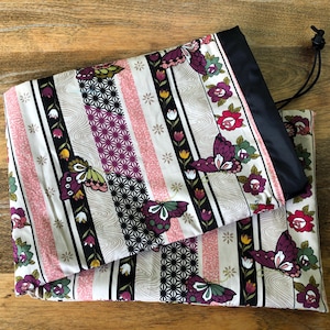 May include: A folded fabric item with a black drawstring. The fabric has a repeating pattern of butterflies, flowers, and geometric shapes in pink, purple, green, and black stripes on a cream background.