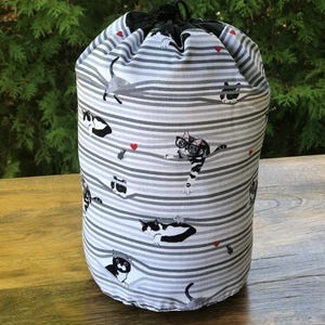 May include: A cylindrical drawstring bag with a gray and white striped pattern. The bag is adorned with illustrations of cats in various poses, some wearing glasses, and small red hearts. The bag is made of fabric and has a black drawstring closure.