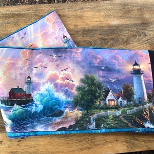 May include: A colourful fabric panel depicting a lighthouse scene. The panel features two lighthouses, one with a red roof and one with a white roof, against a backdrop of a stormy sea and a cloudy sky. The panel has a blue border.