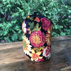 May include: A cylindrical fabric drawstring bag featuring a black background with a vibrant floral pattern of yellow sunflowers, pink and red chrysanthemums, orange lilies, and green leaves. The bag has a black cord closure at the top.