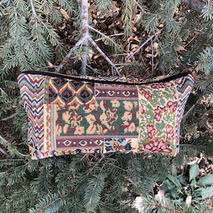 May include: A rectangular tapestry cosmetic bag with a black zipper. The bag features a patchwork design with floral and geometric patterns in shades of green, red, and beige. The bag is resting on a bed of pine needles.