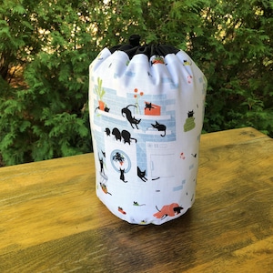 May include: A white drawstring bag with a black top and a whimsical cat print. The fabric features black cats in various poses, set against a light blue brick wall design with potted plants and other decorative elements. The bag is cylindrical and sits on a wooden surface.