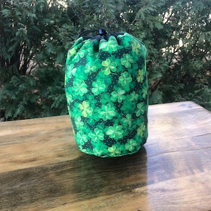 May include: A cylindrical green fabric bag with a drawstring closure. The bag features a pattern of light and dark green shamrocks on a dark green background. The bag is on a wooden surface.