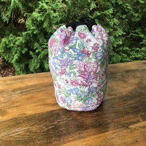 May include: A fabric drawstring bag featuring a floral and fairy design in pink, blue, and purple hues on a white base. It has a black drawstring and is set against a backdrop of green foliage.