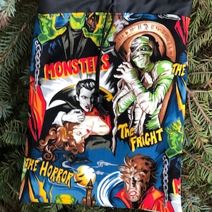 May include: A black drawstring bag with a colorful print of classic monster movie posters. The print features images of Frankenstein, Dracula, the Mummy, and the Wolfman. The text on the posters includes "Monsters", "The Horror", "The Fright", and "The Mummy".