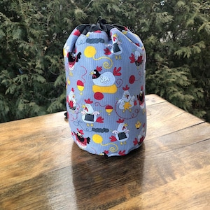 May include: A blue drawstring bag with a repeating pattern of cartoon chickens, yarn balls, and knitting needles. The bag has a black drawstring closure and is sitting on a wooden surface.