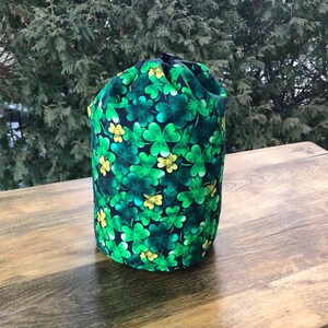 May include: A cylindrical drawstring bag with a pattern of green shamrocks and yellow flowers on a black background. The bag is made of fabric and sits on a wooden surface. The background features green foliage.