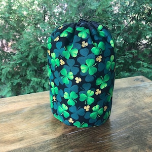 May include: A cylindrical drawstring bag with a black background covered in green and gold shamrocks. The bag is made of fabric and sits on a wooden surface. The bag is a festive item.