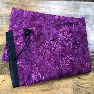 May include: A purple drawstring bag with a floral pattern and black accents. The bag features a large central flower design and smaller swirling patterns. The drawstring is black, and the bag has a black border.