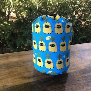 May include: A cylindrical, drawstring bag with a vibrant blue background. The bag is patterned with yellow pineapples, each featuring a black cat face. The bag is sitting on a wooden surface, with a blurred green background.