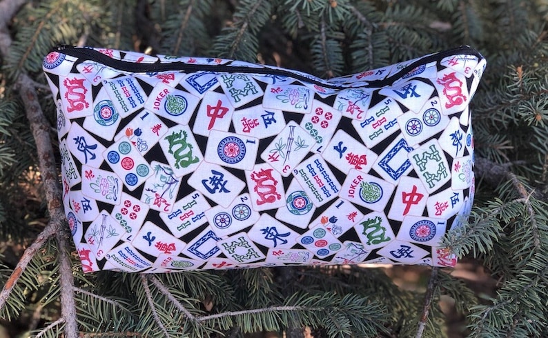 May include: A white zippered pouch with a black background and a pattern of colorful Mahjong tiles. The tiles feature Chinese characters, numbers, and symbols.