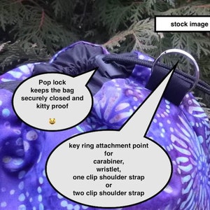 May include: A purple and blue drawstring bag with a pop lock closure. The bag has a key ring attachment point for a carabiner, wristlet, or shoulder strap. The text "Pop lock keeps the bag securely closed and kitty proof" is written on a white speech bubble.