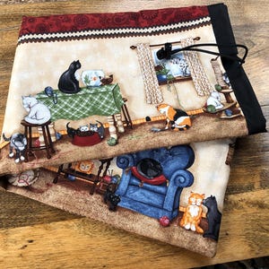 May include: A folded fabric item with a cat-themed design. The pattern shows cats in a domestic setting, with furniture, a green checkered tablecloth, and a window. The top edge has a decorative border and a black elastic cord. The design is reminiscent of a cat's home.