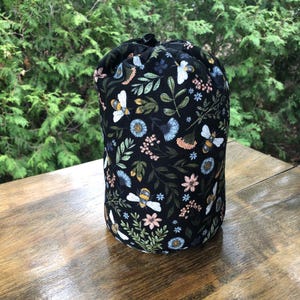 May include: A cylindrical fabric bag with a black background featuring a repeating pattern of yellow and white bees, green leaves, and small blue, pink, and white flowers. The bag has a black drawstring closure at the top opening.