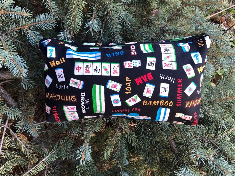 Puede incluir: Bolsa negra con cremallera con un estampado de fichas de Mahjong. La tela presenta varias fichas de Mahjong, dados y caracteres chinos en blanco, rojo, verde y azul. Tambi&eacute;n son visibles las palabras "Mahjong", "Bamboo" y "Summer".