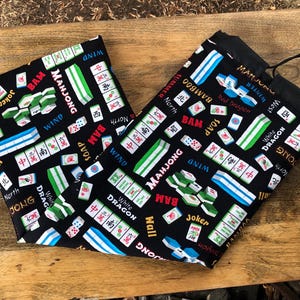 May include: Black drawstring bag with a Mahjong tile pattern. The bag features various Mahjong tiles in green, white, and red, along with text like "Mahjong", "Wind", and "Dragon". The bag is made of fabric.