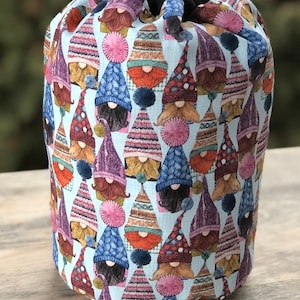 May include: A light blue drawstring bag with a pattern of gnomes wearing colorful knit hats. The gnomes have brown beards and are arranged in a repeating pattern.