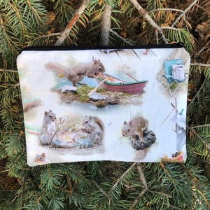 May include: A white zippered pouch with a colorful print of squirrels engaging in various activities, including rowing a boat, sitting by a campfire, and hanging from a tire.